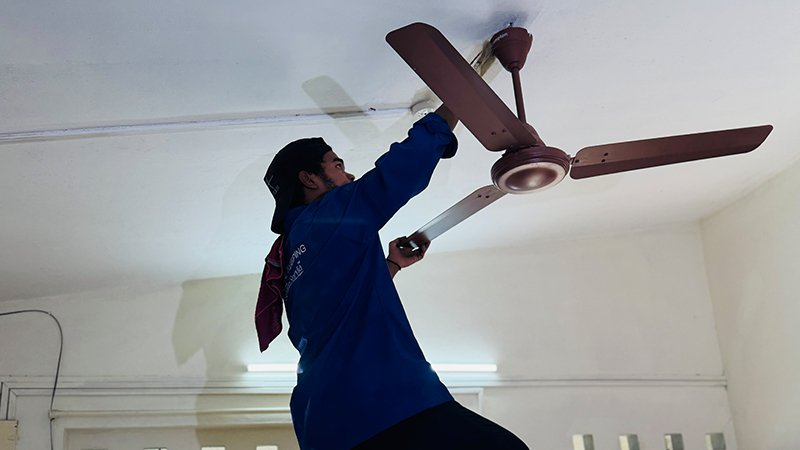 A professional cleaner from Le Servia wearing a blue uniform thoroughly cleaning a ceiling fan, removing dust and ensuring a hygienic living space – part of our deep cleaning service in Kochi.